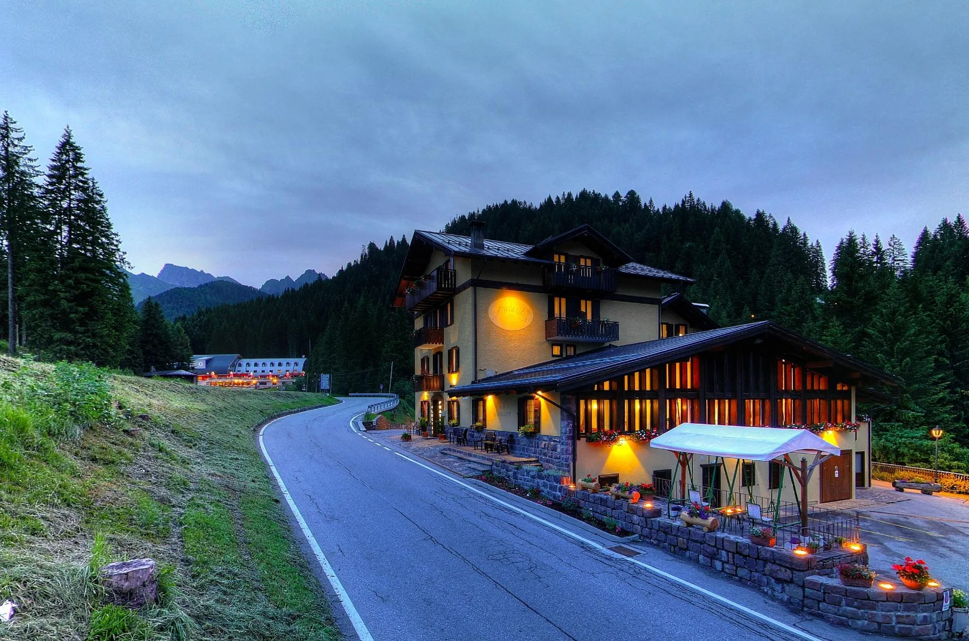 Esterno dell'hotel illuminato al crepuscolo lungo una strada di montagna con dehor e fiori