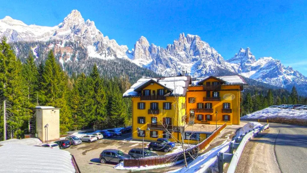 Esterno dell'hotel giallo con parcheggio e Dolomiti innevate sullo sfondo