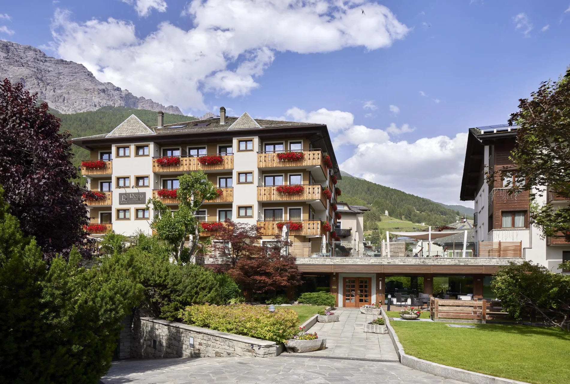 Esterno dell'hotel in estate con balconi fioriti, giardino verde e montagne
