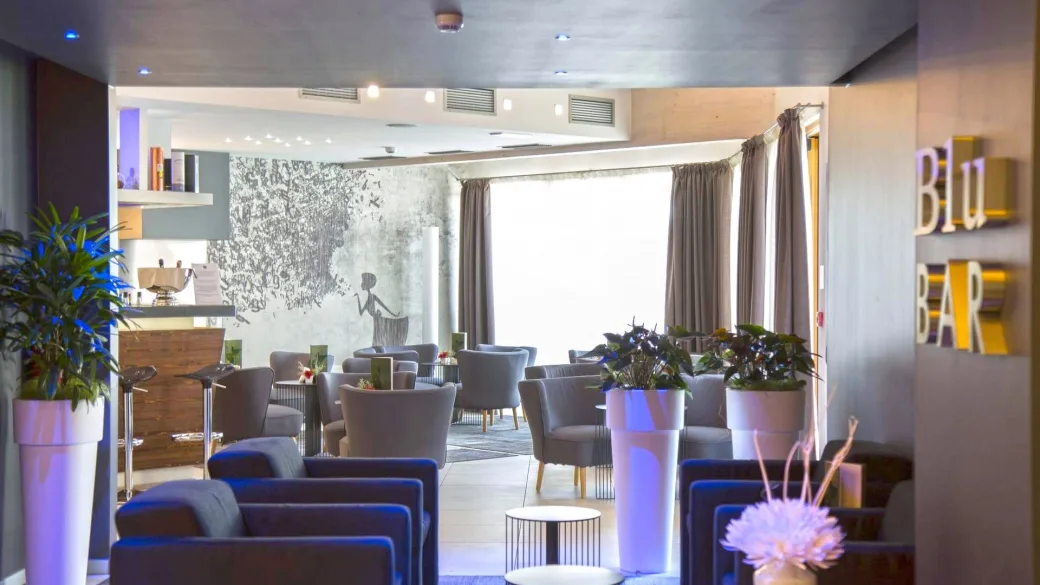 Lounge bar dell'hotel con poltrone grigie, piante decorative e insegna Blu Bar