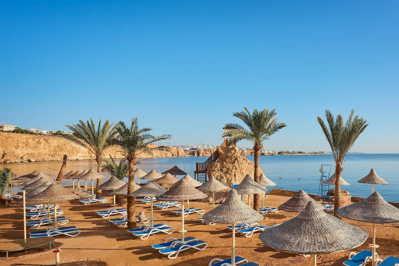Spiaggia con ombrelloni di paglia, palme e lettini blu sulla costa rocciosa