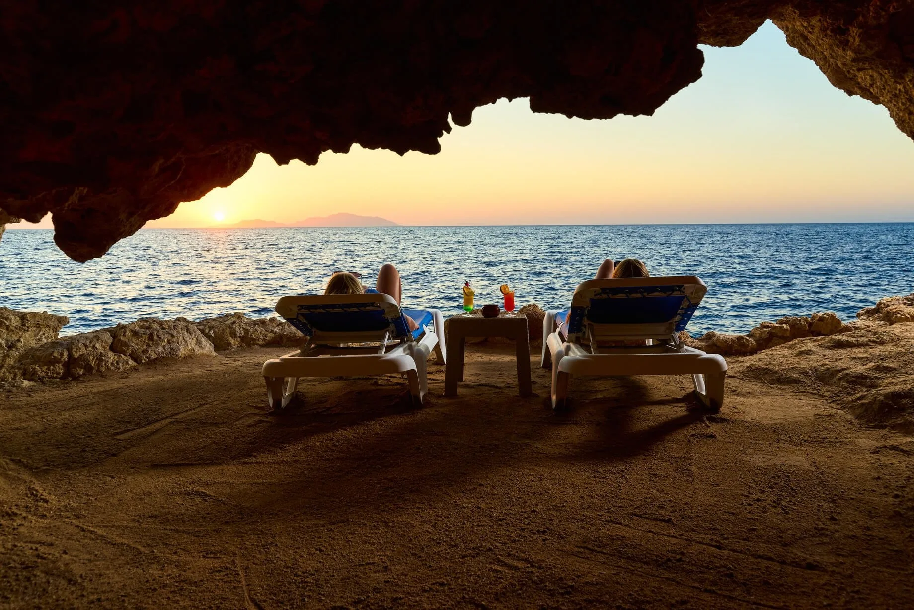 Coppia rilassata su lettini davanti a una grotta ammirando il tramonto sul mare