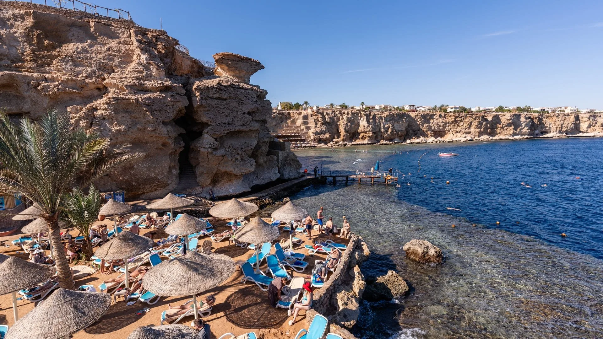 Baia con spiaggia attrezzata tra scogliere e persone che fanno snorkeling nel mare limpido