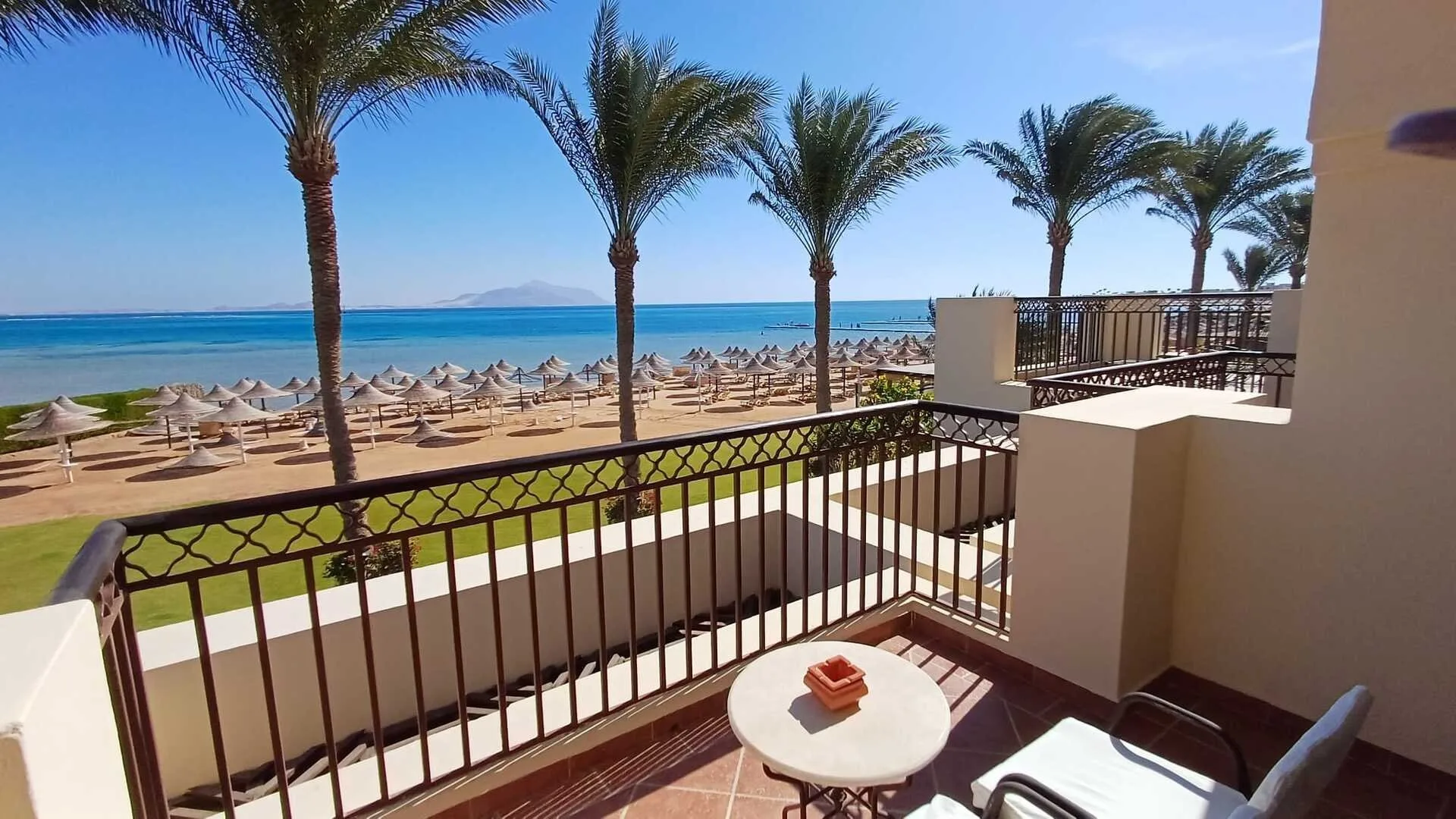 Balcone con vista panoramica sul mare, palme, spiaggia con ombrelloni e tavolino