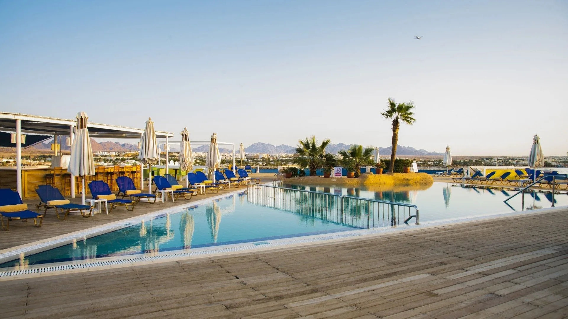 Grande piscina su terrazza in legno con lettini blu, palme e vista sul deserto montuoso