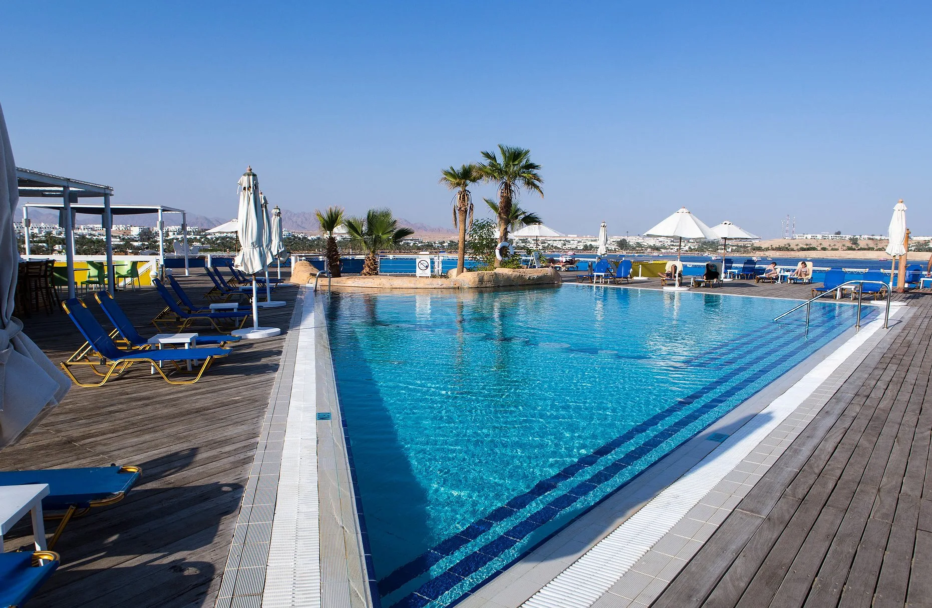 Piscina azzurra con lettini e ombrelloni su deck in legno affacciata sulla baia del Mar Rosso