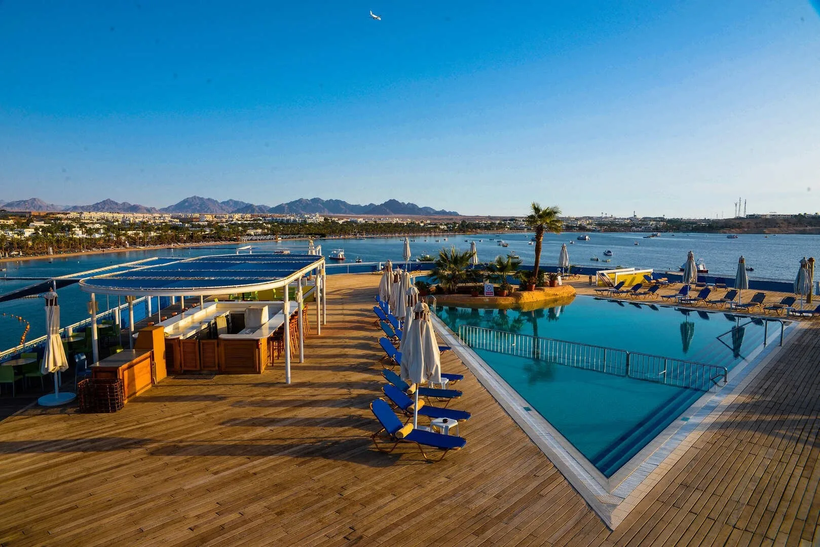 Terrazza in legno con piscina, lettini blu e bar affacciata sul mare e sulle montagne