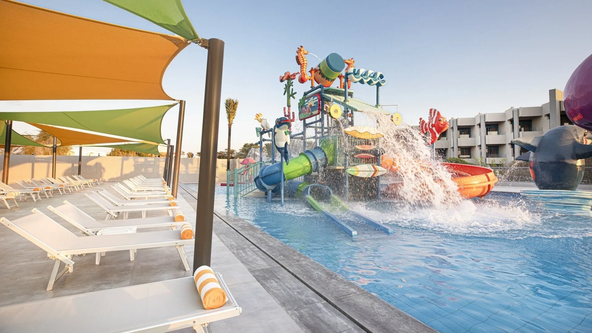Parco acquatico per bambini con giochi d'acqua e scivoli colorati, lettini e teli sotto tende colorate