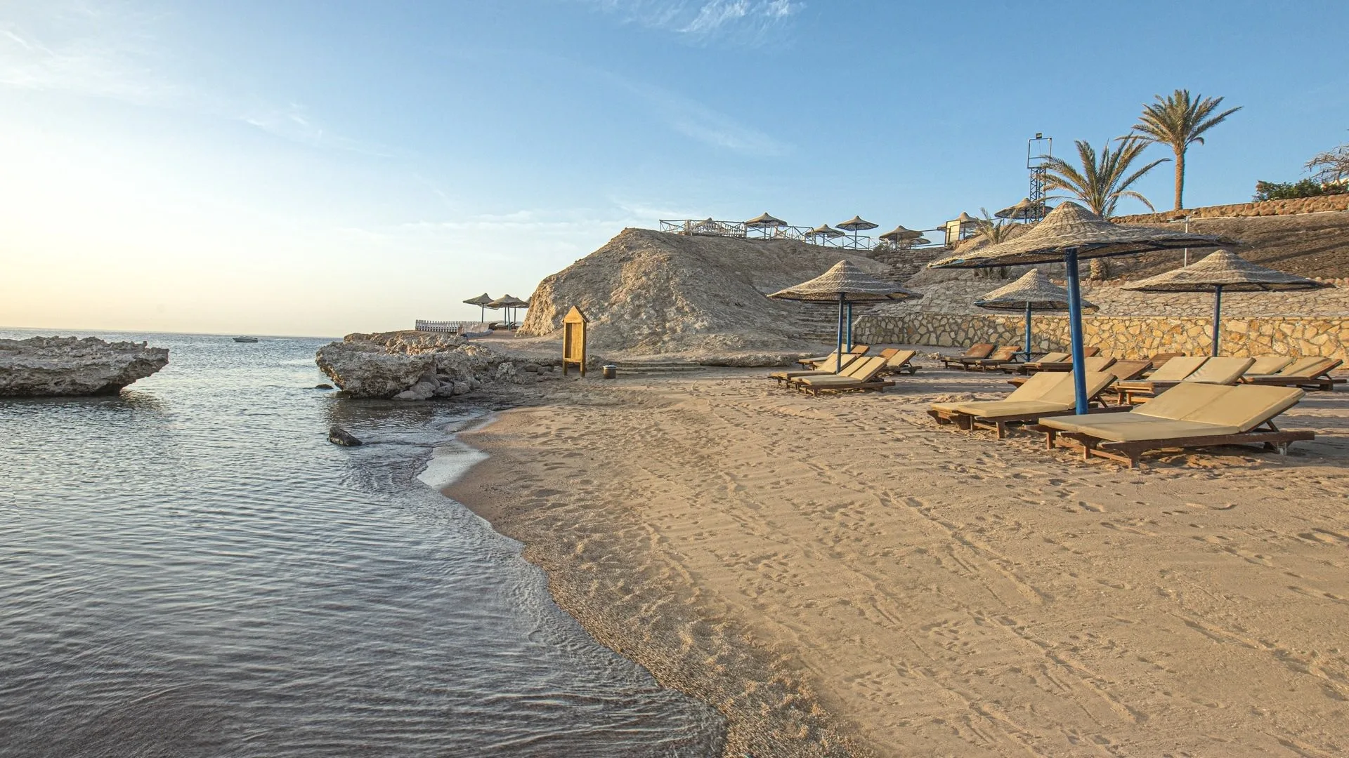 Spiaggia sabbiosa con lettini e ombrelloni in paglia affacciata su una baia rocciosa al tramonto