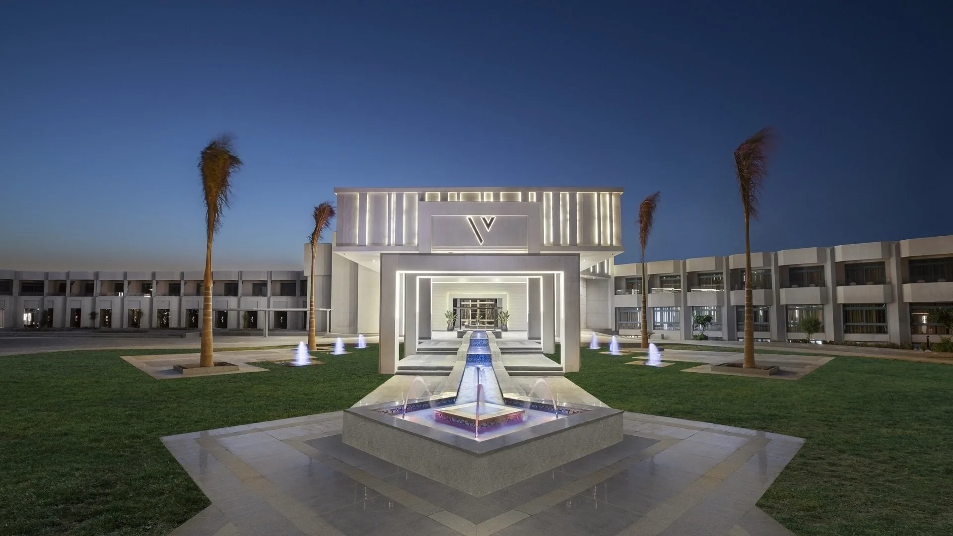 Ingresso illuminato del resort con fontane, palme e prato verde di sera