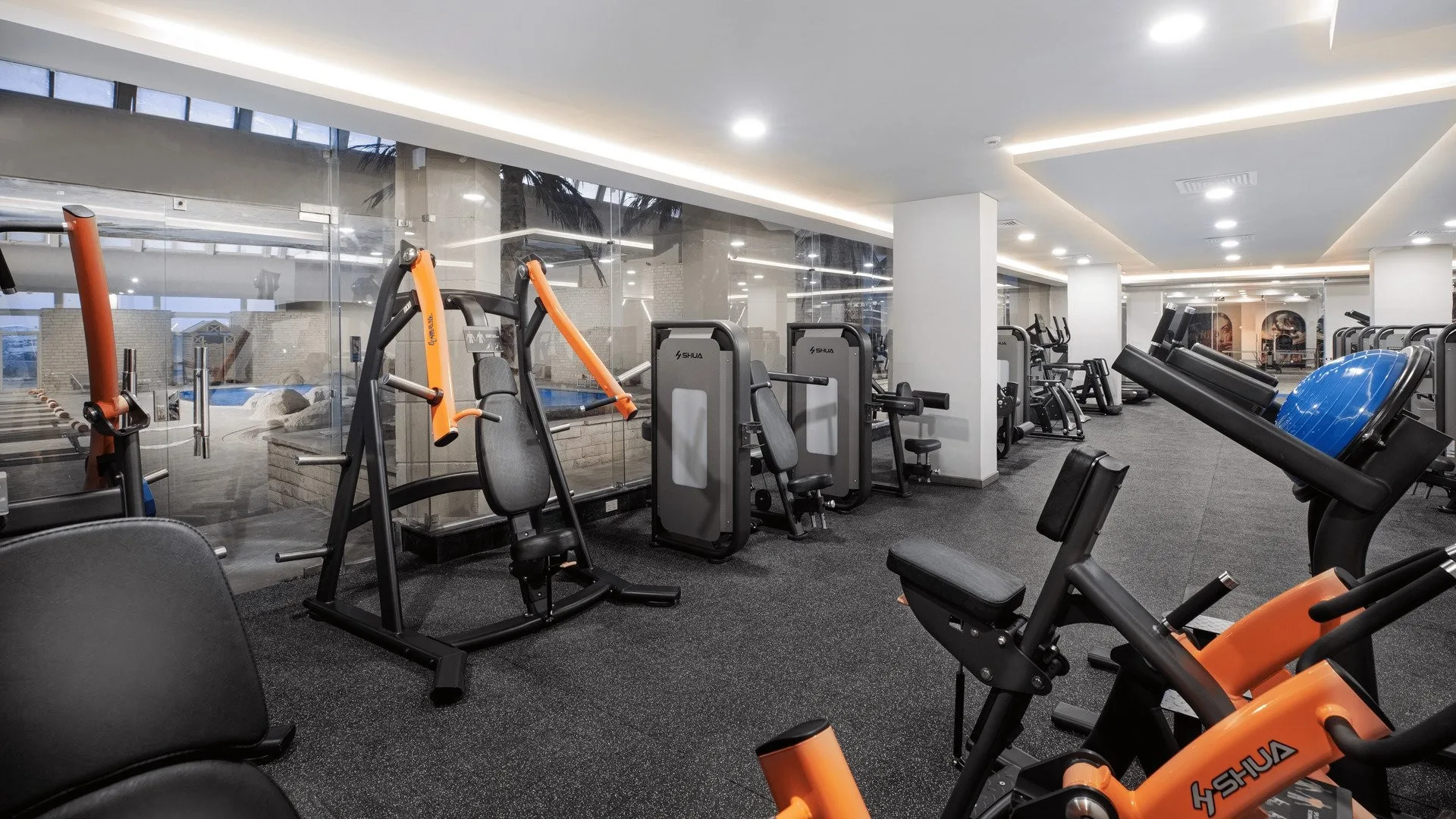Palestra attrezzata con macchinari arancioni e cardio, affacciata su piscina interna