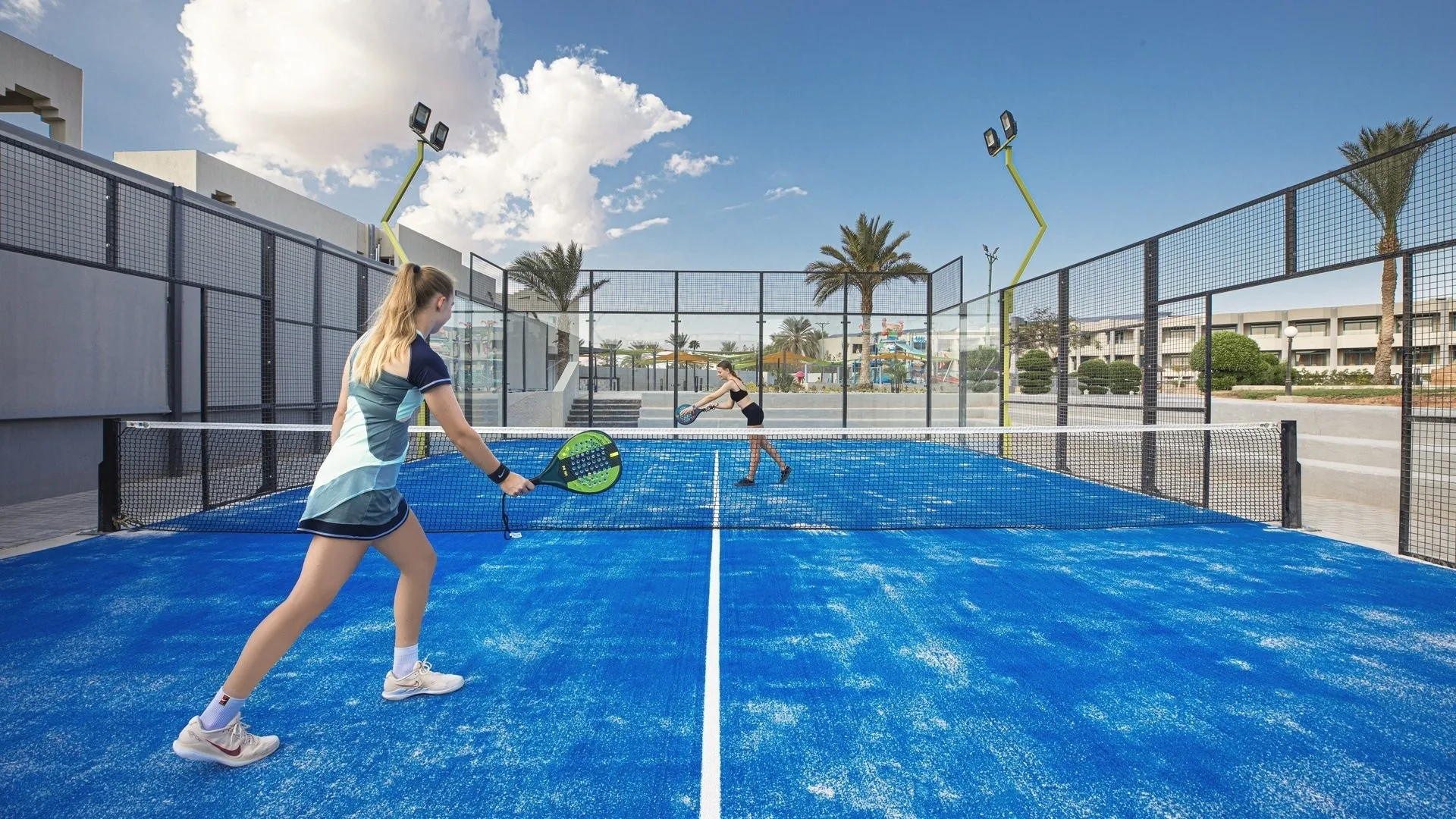 Due persone giocano a padel su campo blu all'aperto con palme sullo sfondo