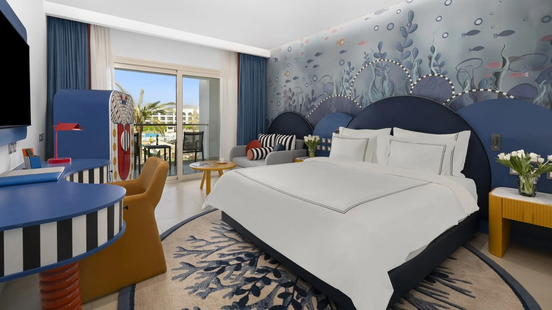 Camera a tema marino con letto matrimoniale, parete decorata con fondale oceanico, arredi colorati e balcone affacciato sulla piscina