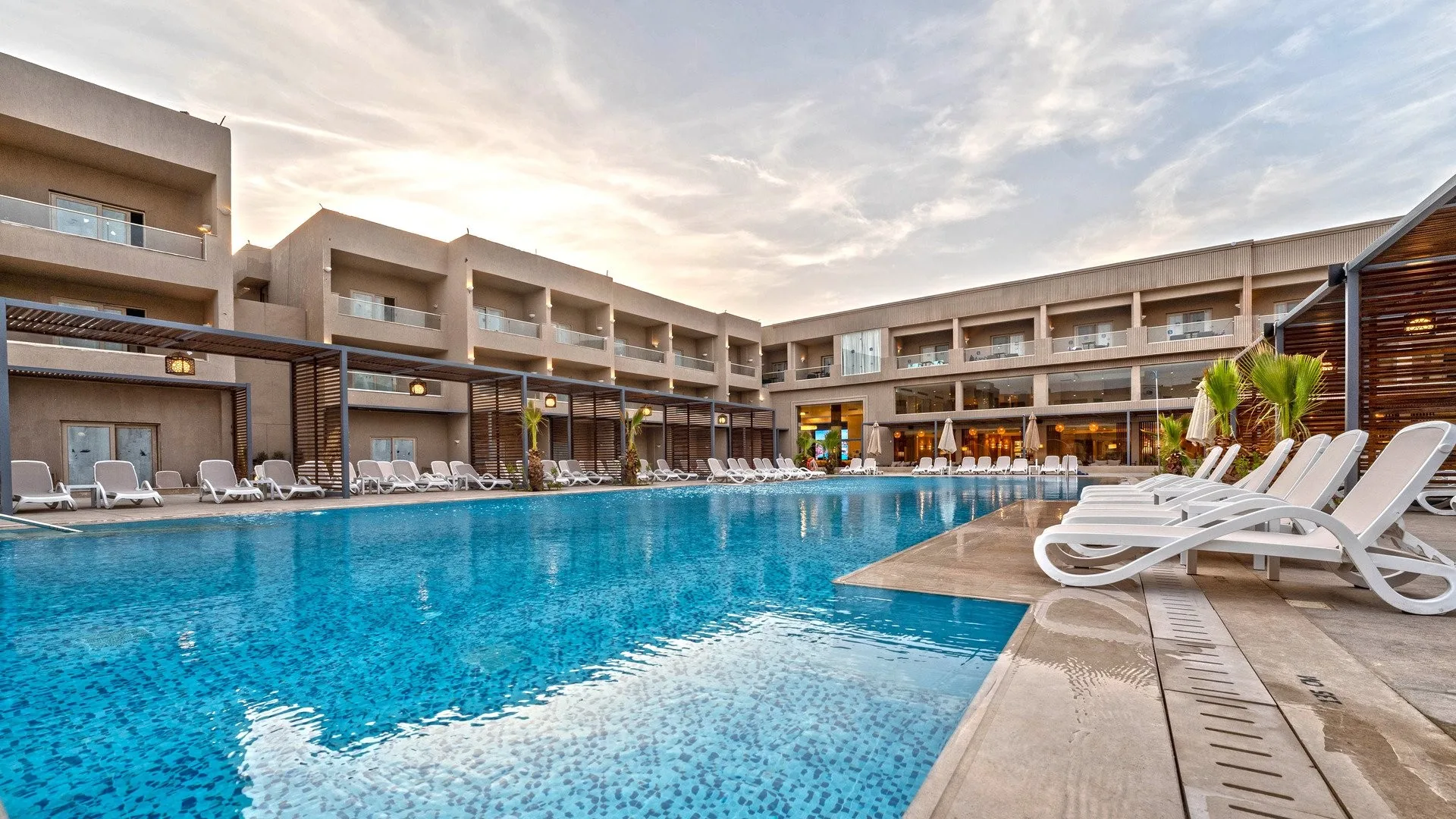 Piscina rettangolare con lettini bianchi circondata dagli edifici del resort al tramonto