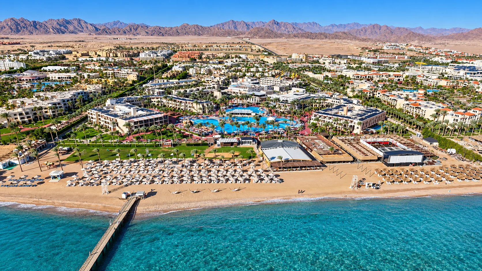 Vista aerea del resort sulla spiaggia con piscine, ombrelloni e montagne desertiche sullo sfondo
