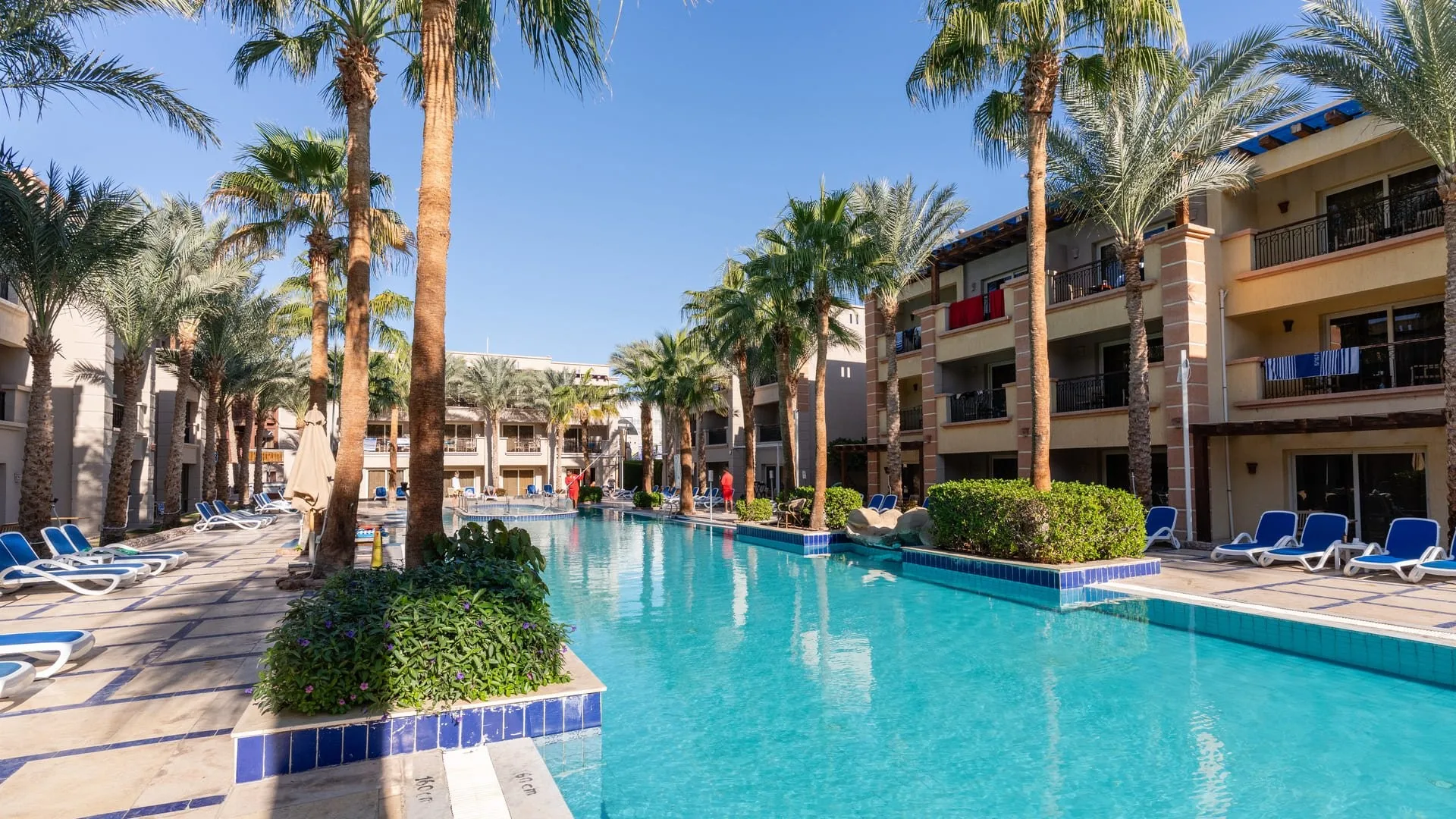 Piscina tra gli edifici del resort con palme, lettini bianchi e blu