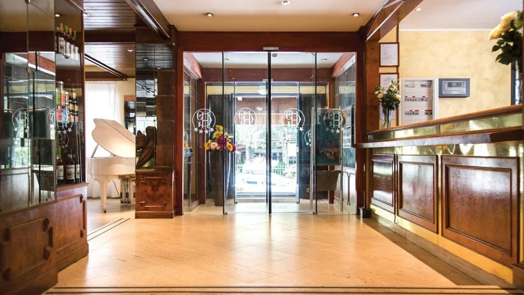 Reception e ingresso dell'hotel con porta a vetri, pavimento in legno e pianoforte bianco