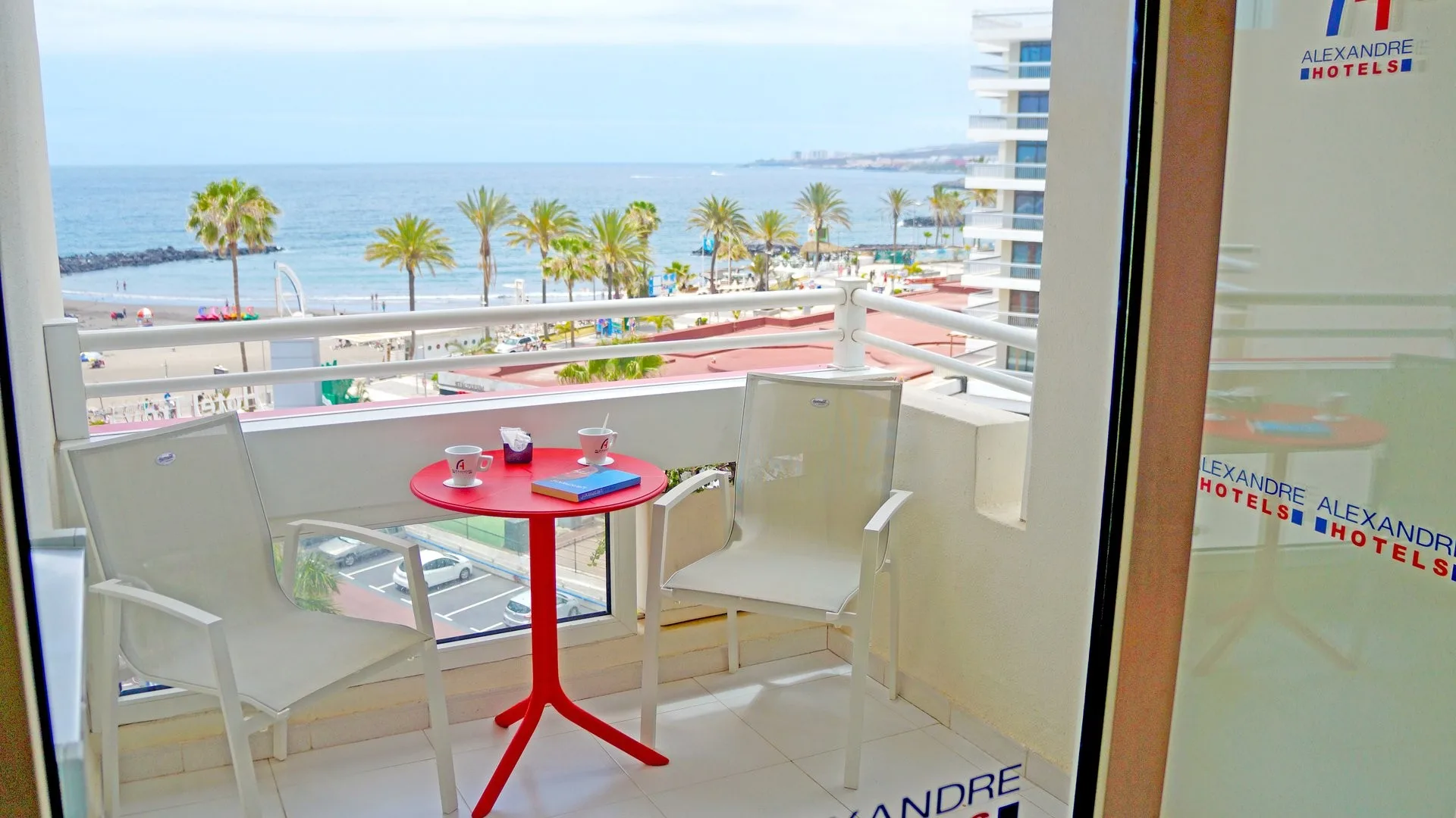 Balcone con tavolino rosso e sedie bianche affacciato sul mare e la spiaggia