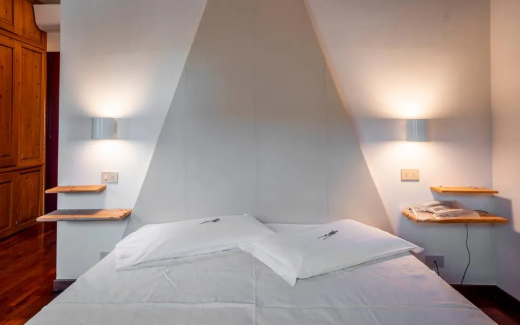Dettaglio di letto con mensole in legno, applique e parete triangolare bianca