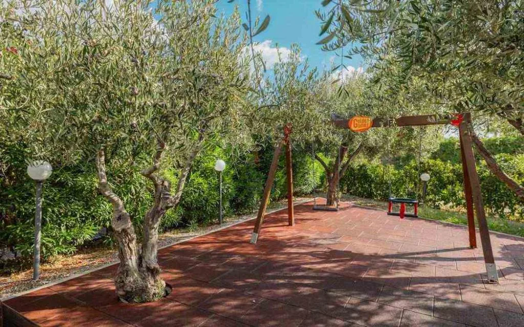 Area giochi nel giardino con altalene tra ulivi e vialetto in piastrelle rosse