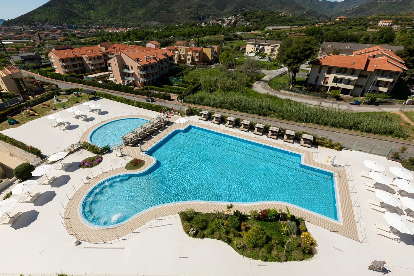 Vista aerea della piscina a forma sinuosa del resort con lettini, ombrelloni e panorama sulle montagne