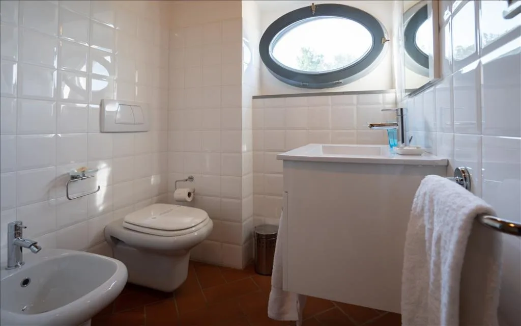 Bagno luminoso con piastrelle bianche, WC, bidet, lavabo e finestra ovale