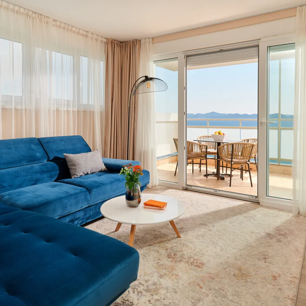 Soggiorno con divano blu, tavolino bianco e terrazza con tavolo e vista mare
