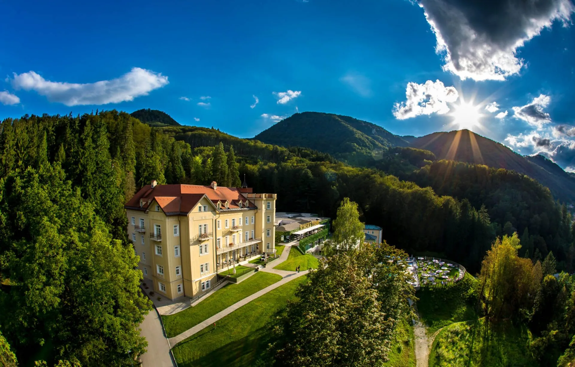 Hotel storico color crema tra le colline boscose con cielo azzurro e sole