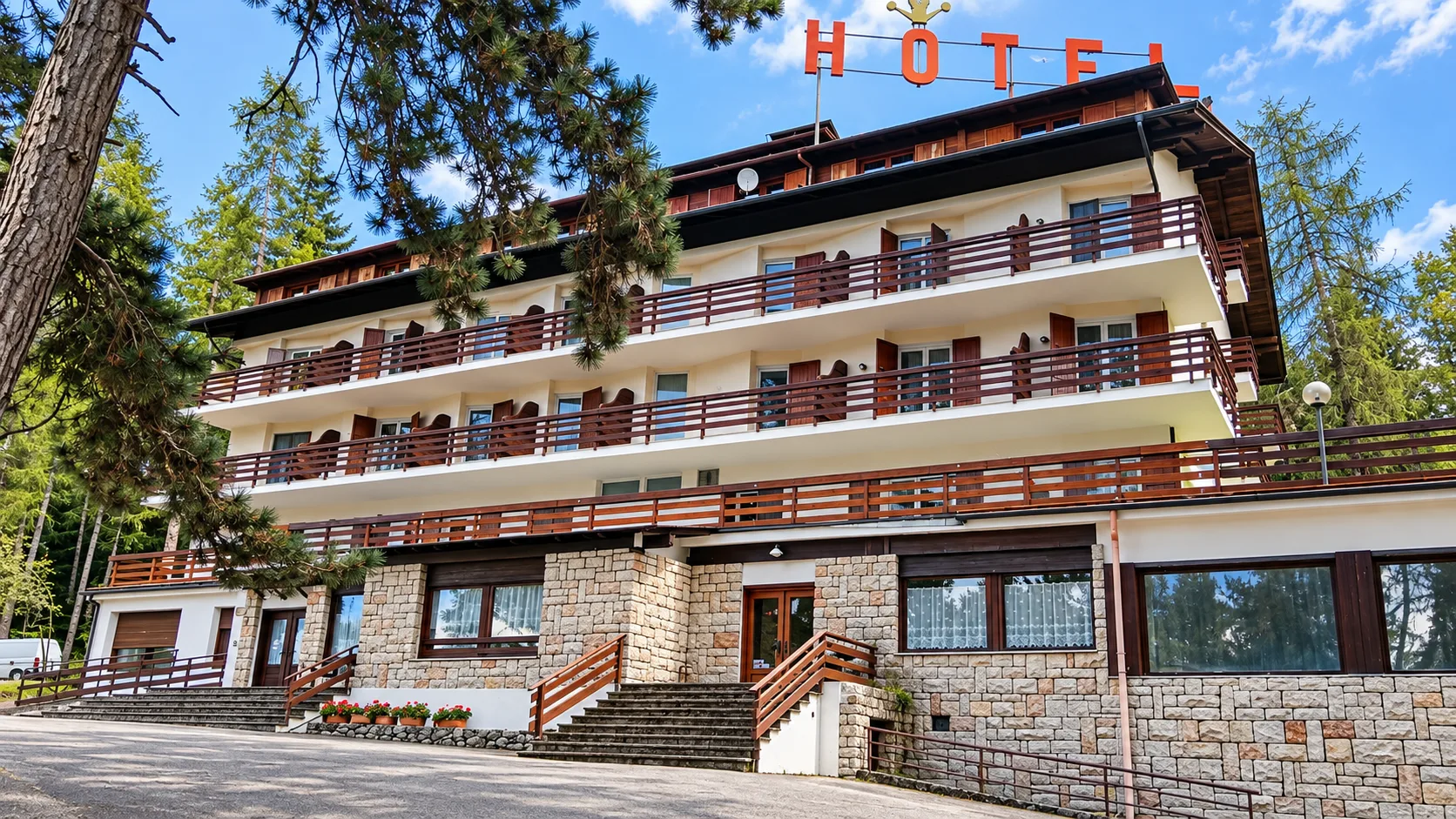 Facciata dell'hotel con balconi in legno, insegna HOTEL e base in pietra tra i pini
