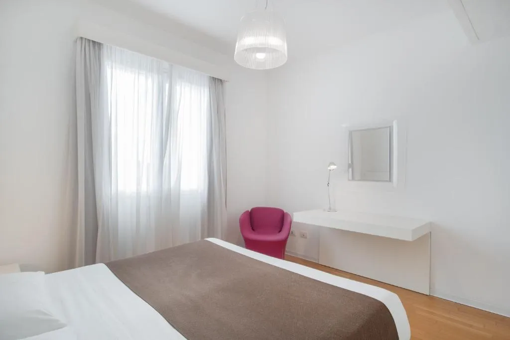 Camera da letto bianca con finestra luminosa, scrivania e poltrona fucsia