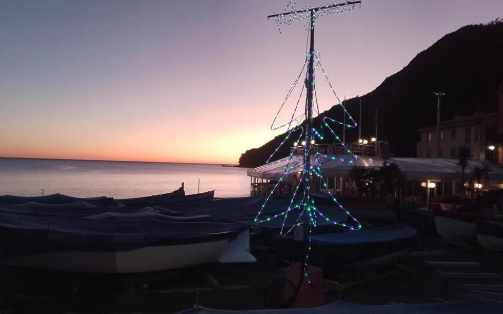 Albero di Natale luminoso sulla spiaggia con barche al tramonto e mare sullo sfondo