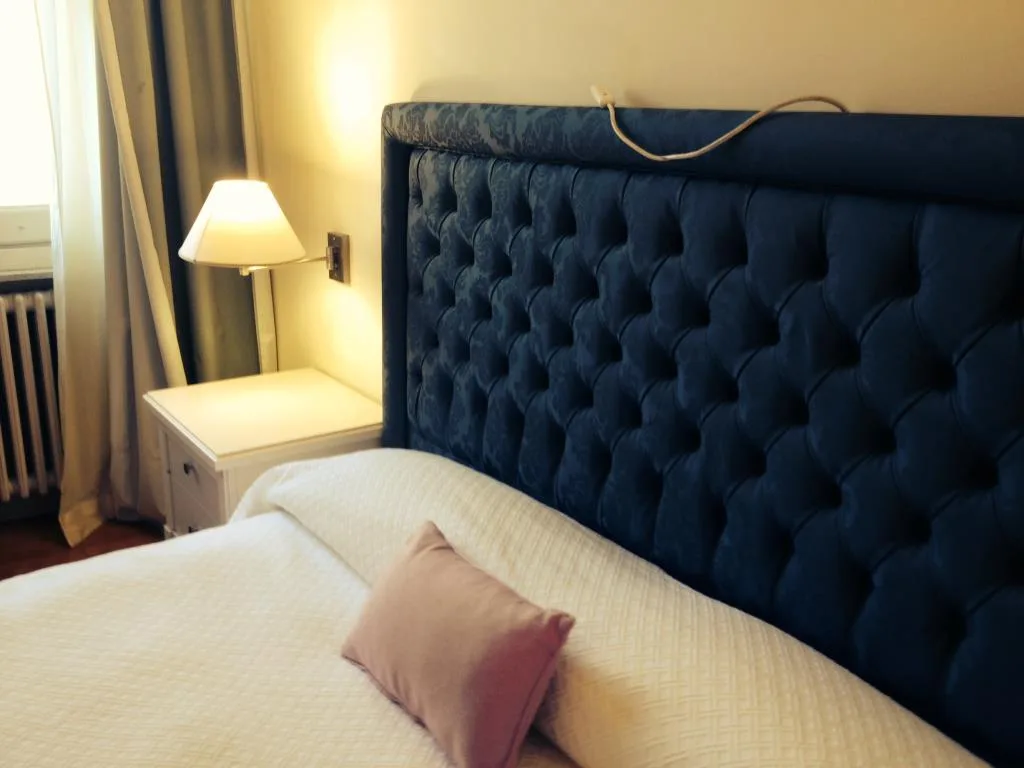 Dettaglio di letto con testiera blu capitonné, lampada accesa e cuscino rosa