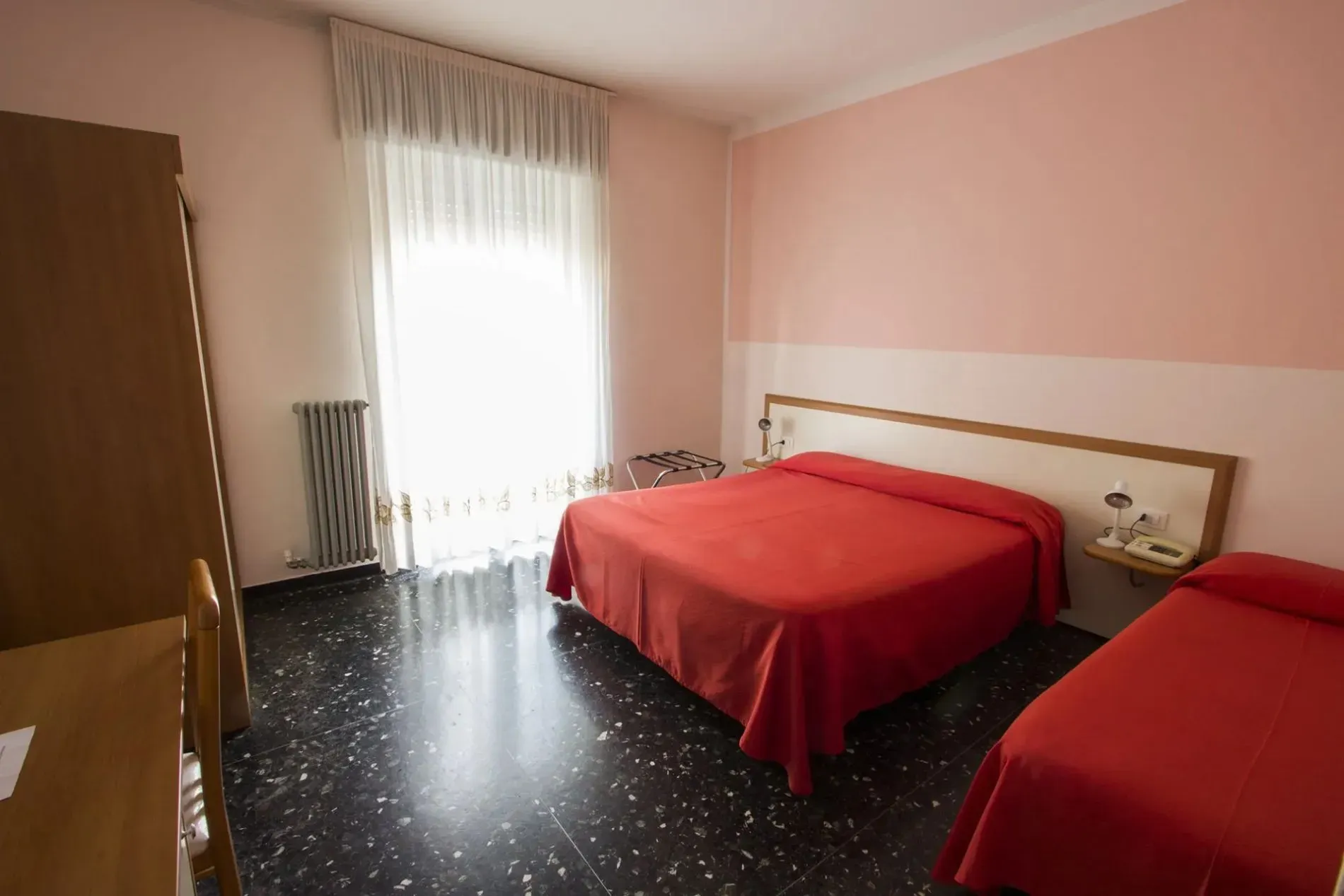 Camera tripla con letti dalle coperte rosse, pareti rosa e grande finestra luminosa