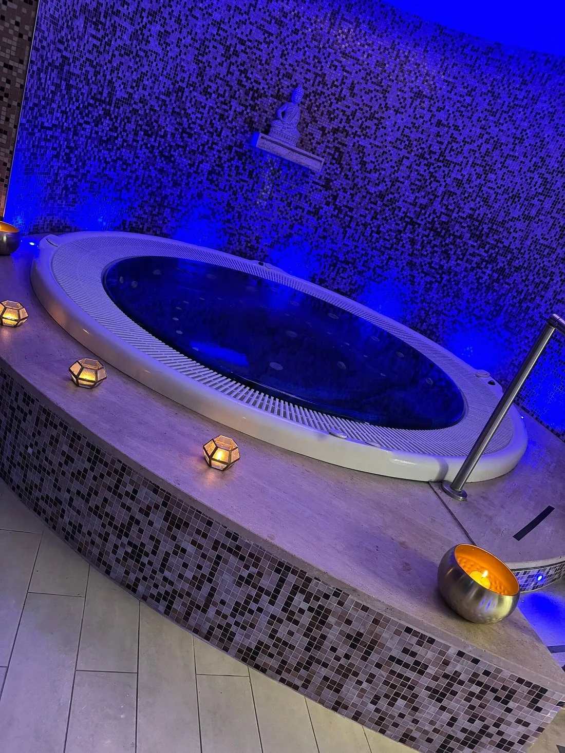 Vasca idromassaggio ovale con illuminazione blu e candele su parete a mosaico