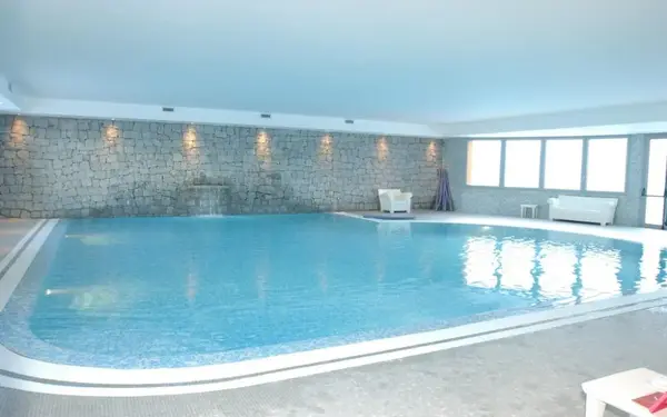 Piscina coperta con acqua azzurra, cascata e parete in pietra con poltrone bianche