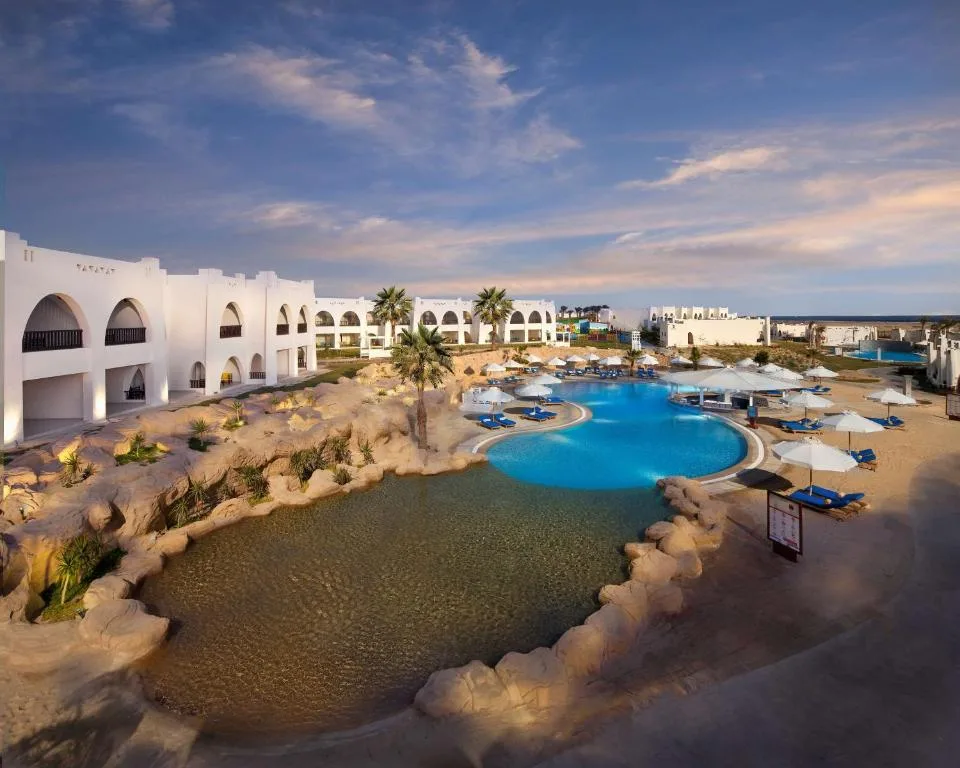 Vista aerea del resort con piscina blu, laghetto naturale roccioso e edifici bianchi al tramonto