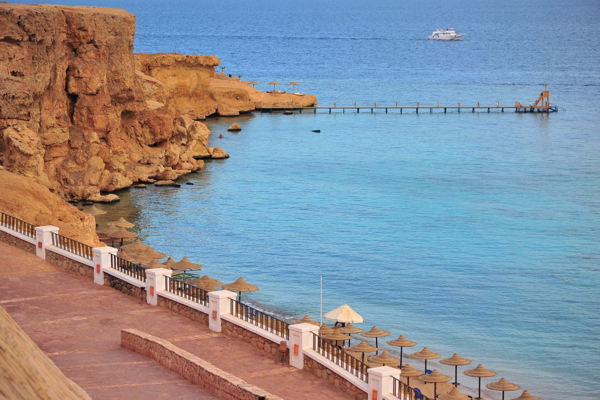 Baia rocciosa con pontile che si estende sul mare turchese e ombrelloni in paglia lungo la costa