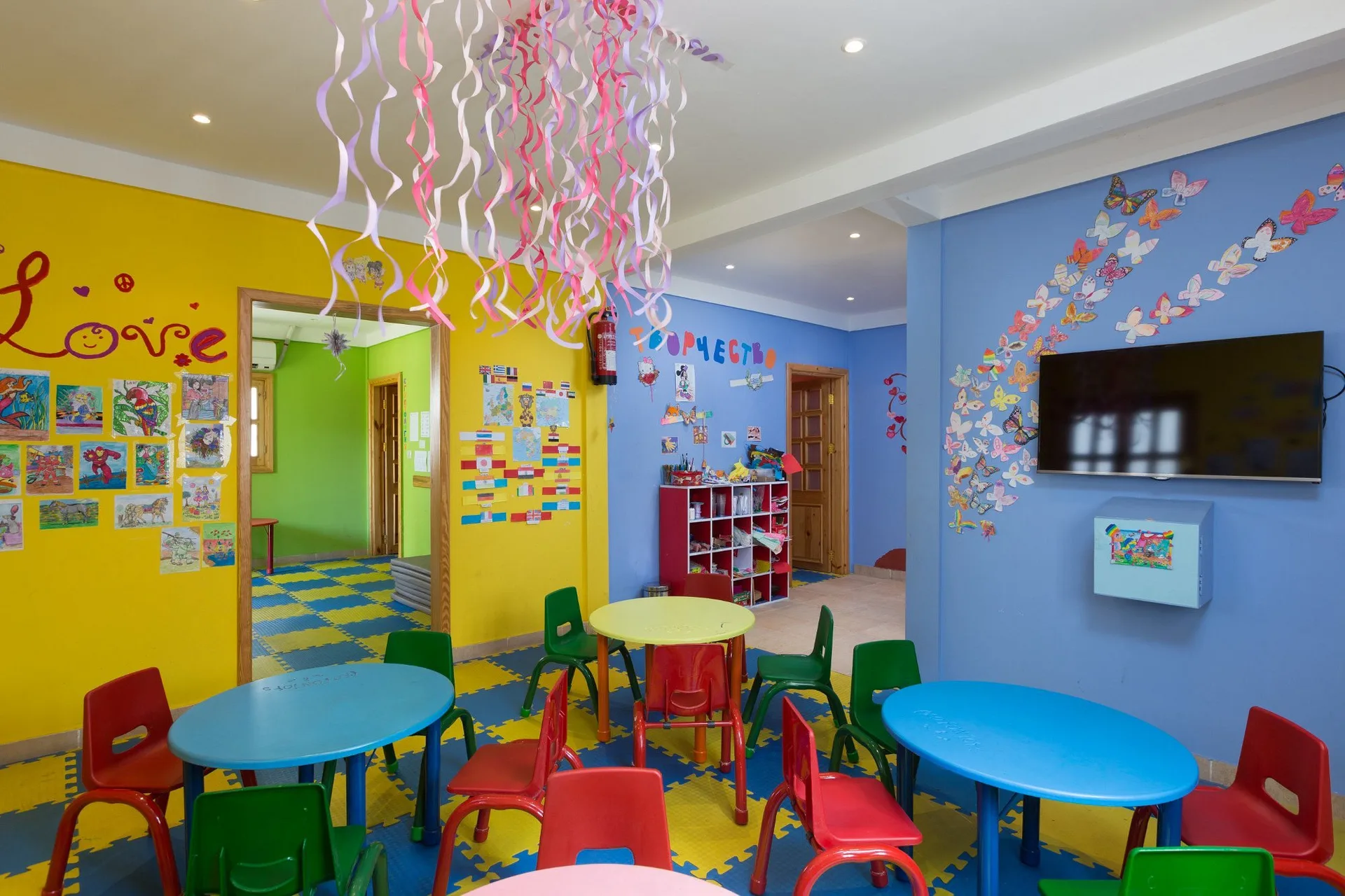 Sala giochi per bambini colorata con tavolini, sedie e TV a parete