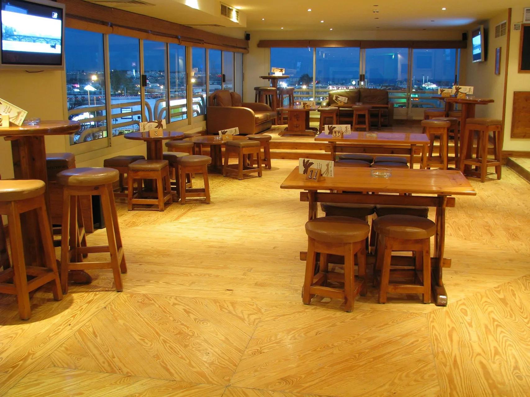 Interno di un bar con tavoli e sgabelli in legno, pavimento chiaro e vista notturna