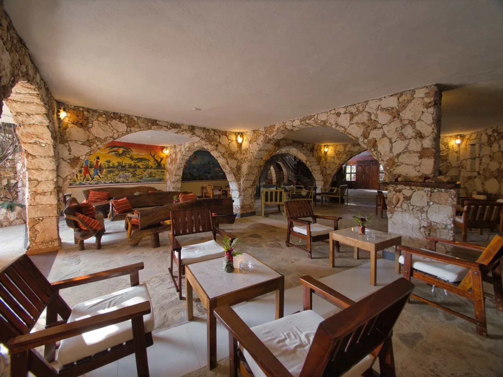 Sala interna con pareti e archi in pietra, poltrone in legno con cuscini chiari e murales colorati