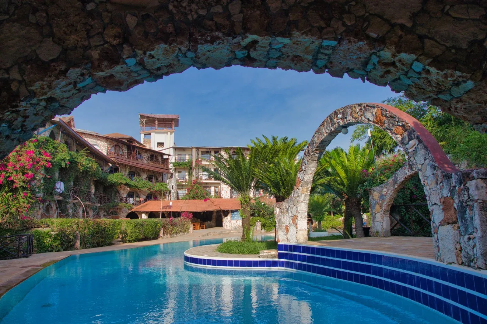 Piscina del resort vista attraverso un'arcata in pietra con palme e edificio sullo sfondo