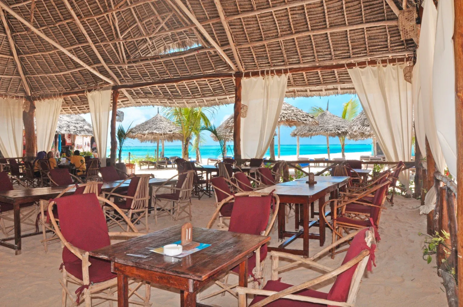Ristorante sulla spiaggia con tetto in paglia, tavoli in legno e vista sull'oceano turchese